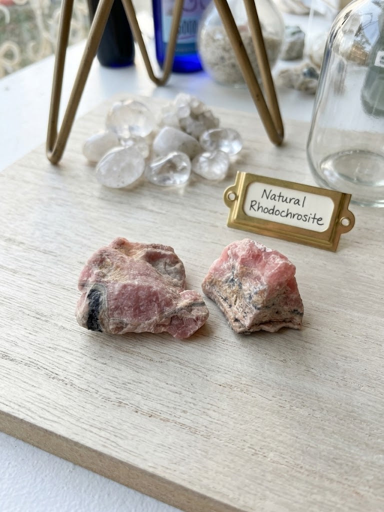A detailed macro close-up photograph of two raw Rhodochrosite rough stones, highlighting their intricate crystal banding, varying rose-pink colors, and black manganese oxide matrix inclusions.[cite: 1,cite: 1]