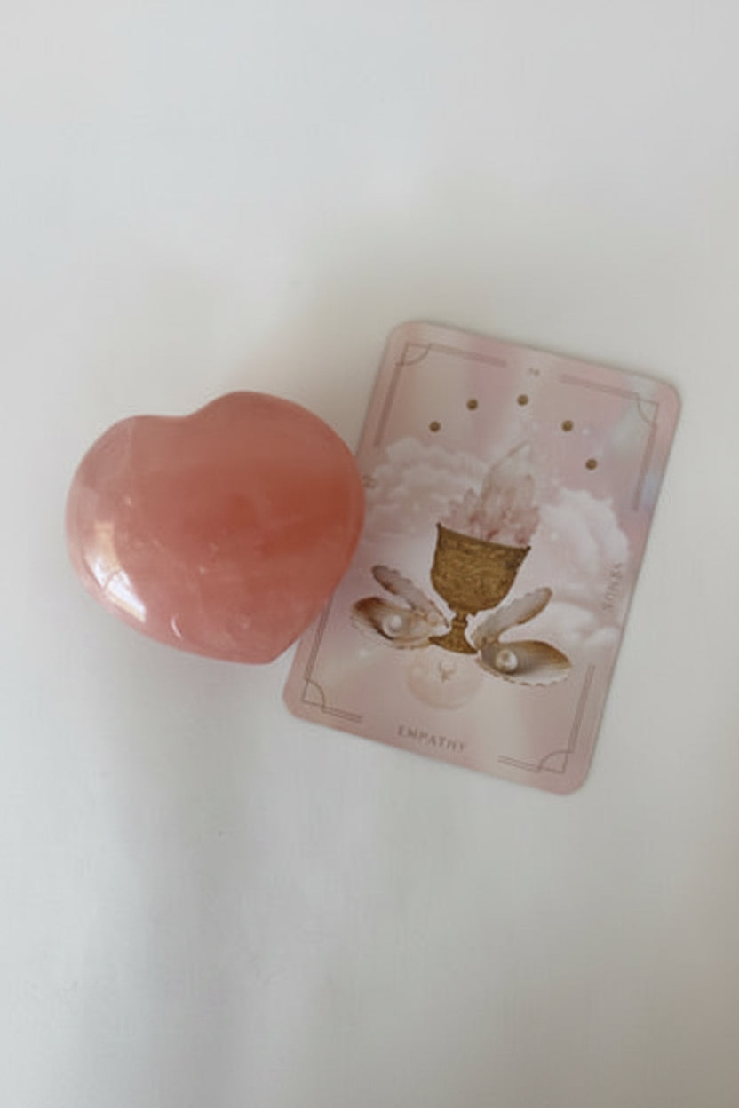 A polished pink heart-shaped crystal resting on top of a small illustrated tarot card on a plain light background.