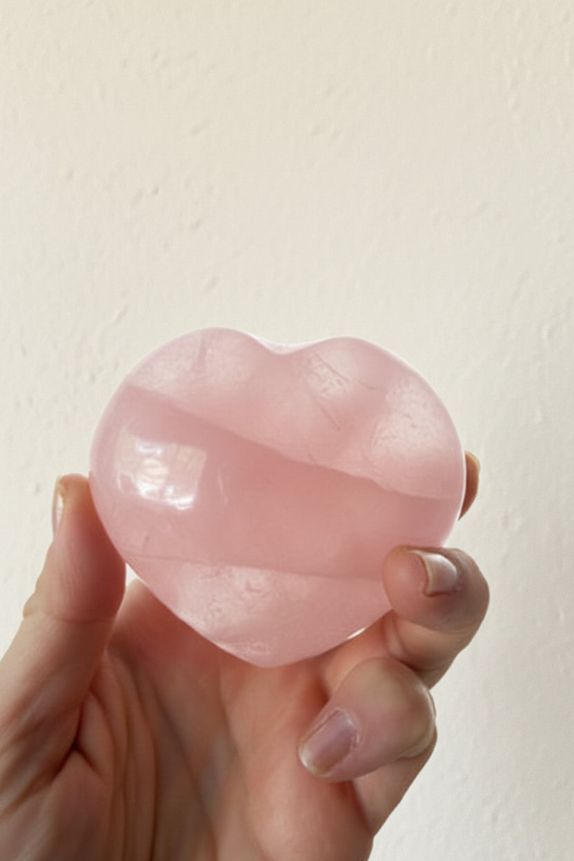 A hand holding a smooth, polished pink heart-shaped stone against a plain light background.