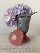 A smooth, polished pink rose quartz crystal sphere in the foreground, with a blue-gray ceramic vase holding light purple hydrangea flowers behind it on a beige textured fabric surface.