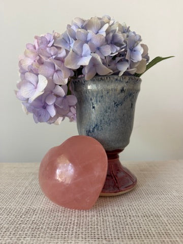 A smooth, polished pink rose quartz crystal sphere in the foreground, with a blue-gray ceramic vase holding light purple hydrangea flowers behind it on a beige textured fabric surface.